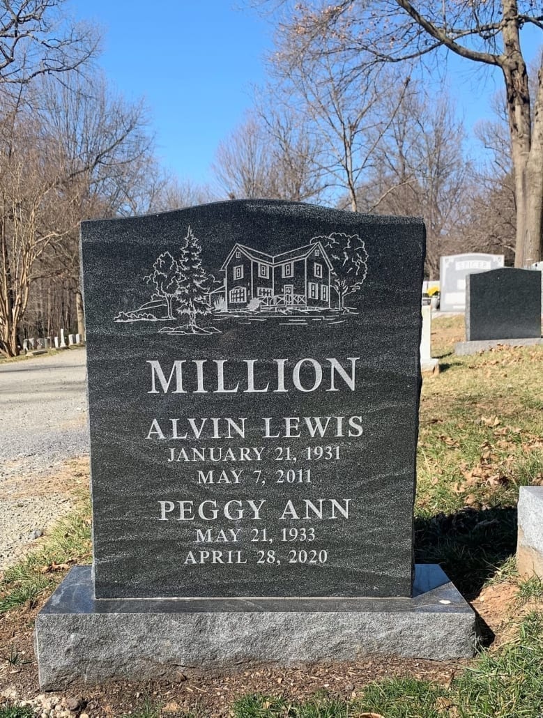 Dark granite upright monument etched with a farmhouse and trees, standing in a cemetery